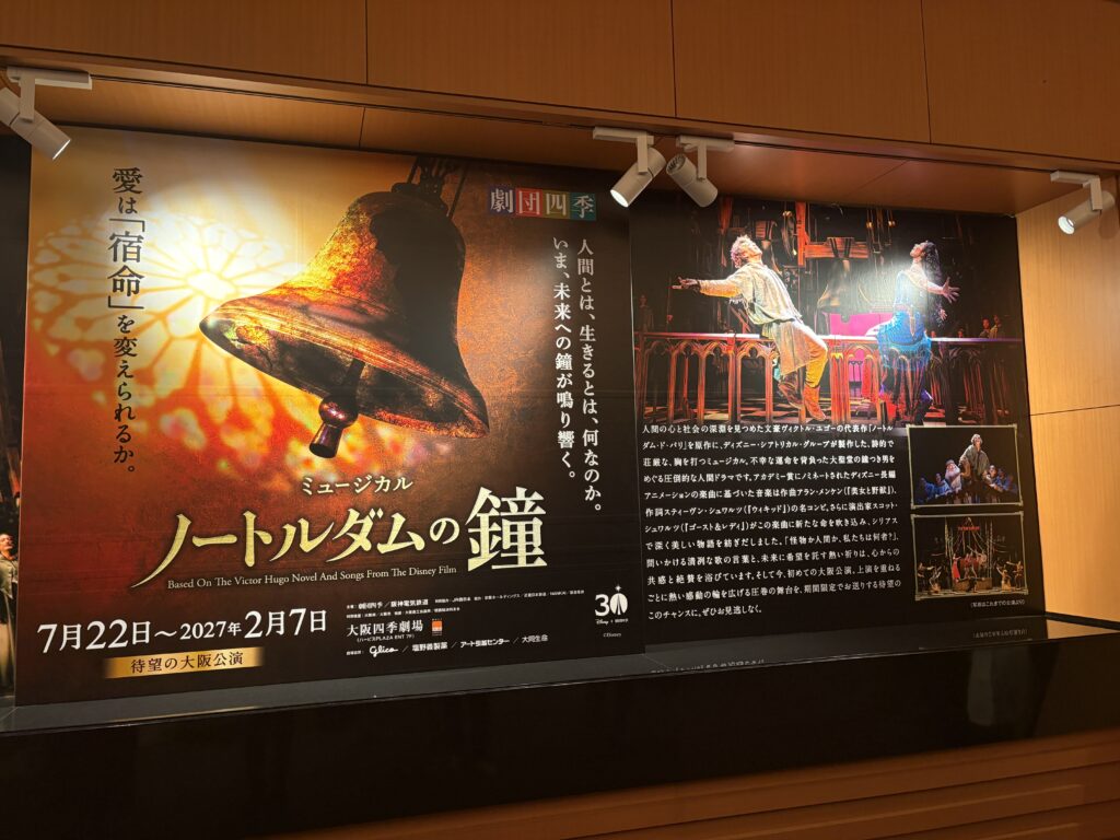 Large promotional display for Shiki Theatre's The Hunchback of Notre Dame musical in Osaka, featuring a golden cathedral bell and photographs of the stage production.