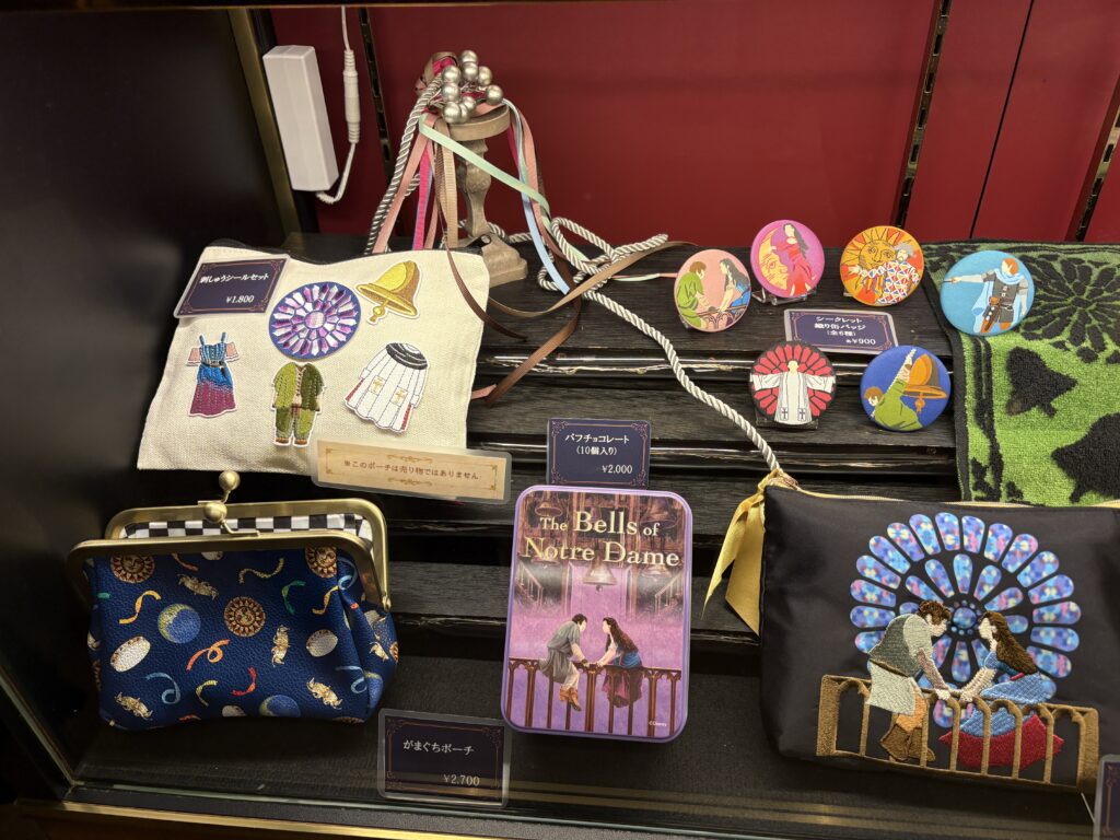The Hunchback of Notre Dame musical merchandise displayed at Shiki Theatre Osaka, including embroidered pouches, a tote bag, collectible badges, and themed accessories.