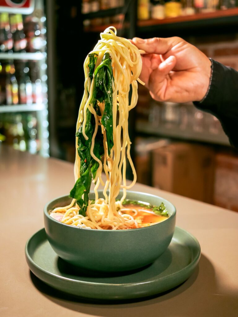 Noodles and leafy greens being lifted from a bowl with chopsticks.