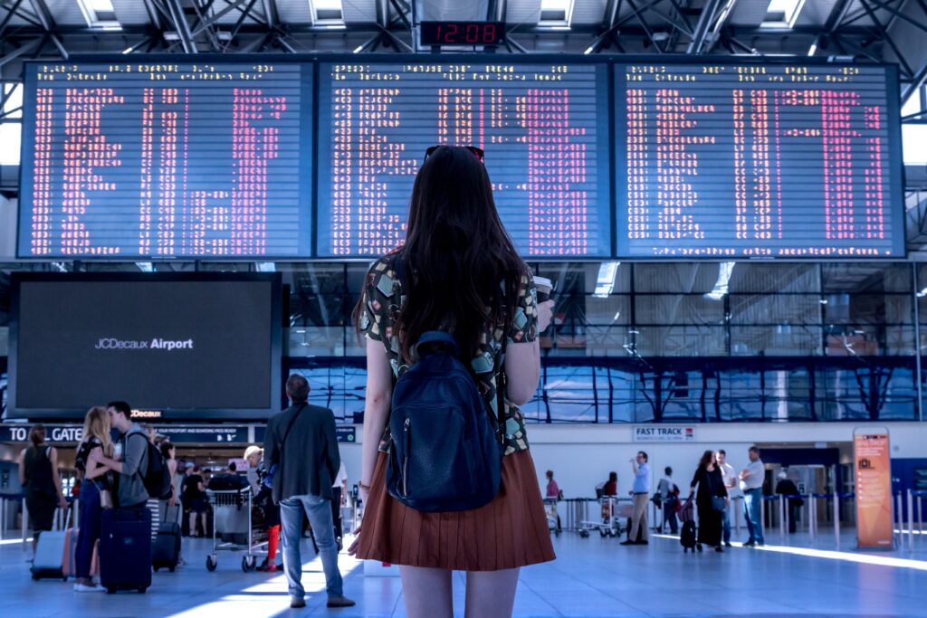 Traveler with a backpack looking at flight information boards inside a busy airport terminal.