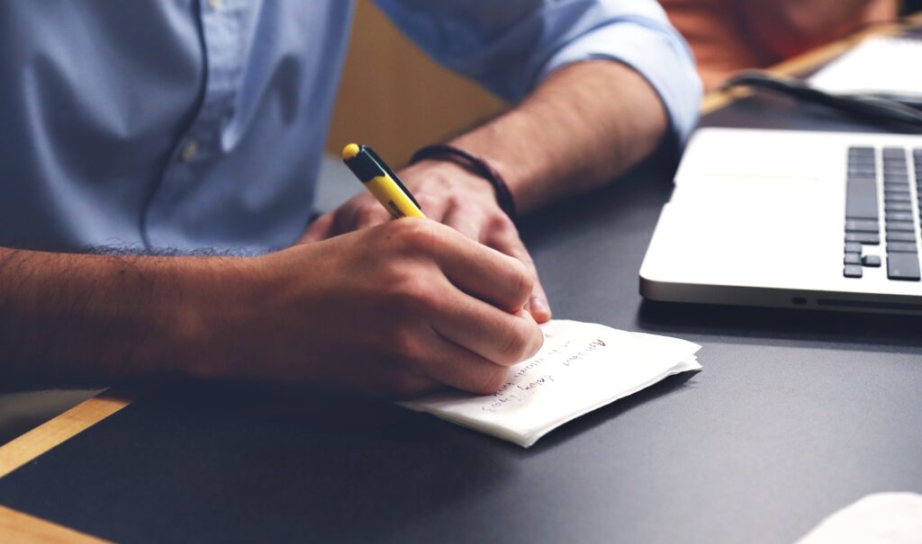 A person writing notes beside a laptop while preparing documents for a job application.