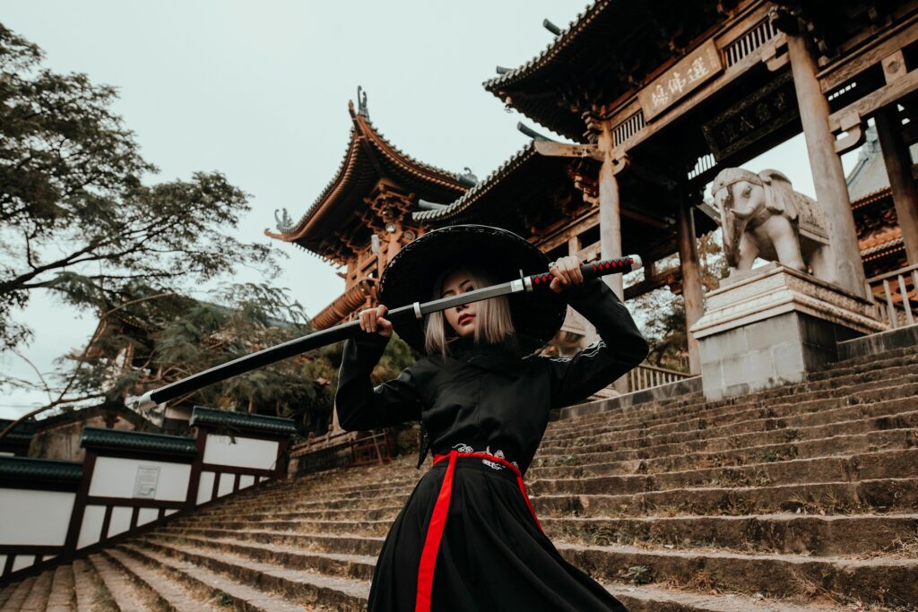 A person dressed in a black samurai-inspired costume holds a sword in front of a traditional Japanese-style building.