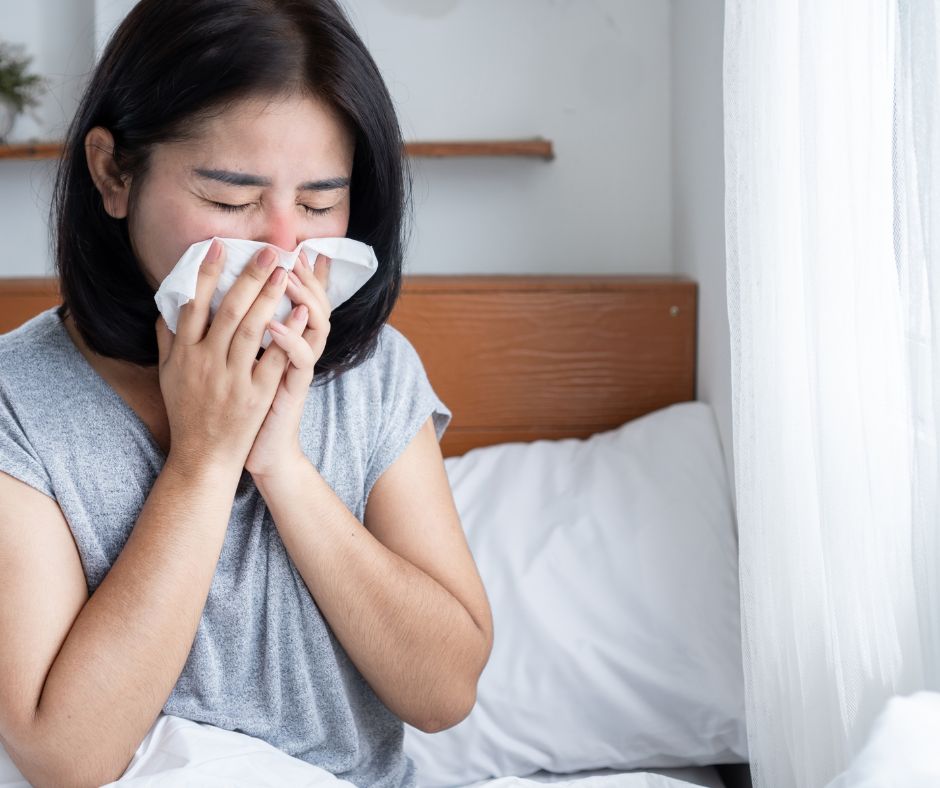 Woman sneezing into a tissue while sitting on a bed near a window.