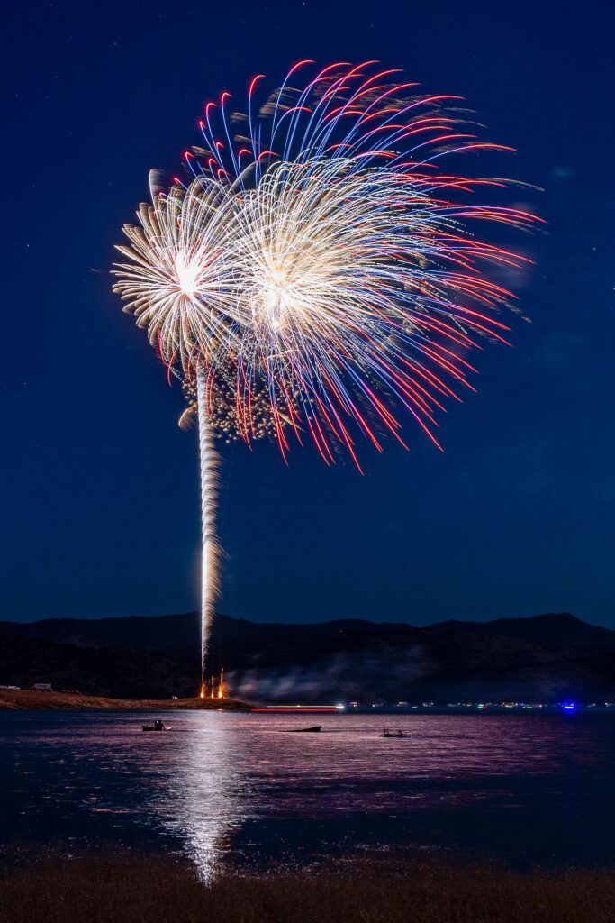 Large red, blue, and white fireworks burst over a lake at night, illuminating the water and distant mountains.