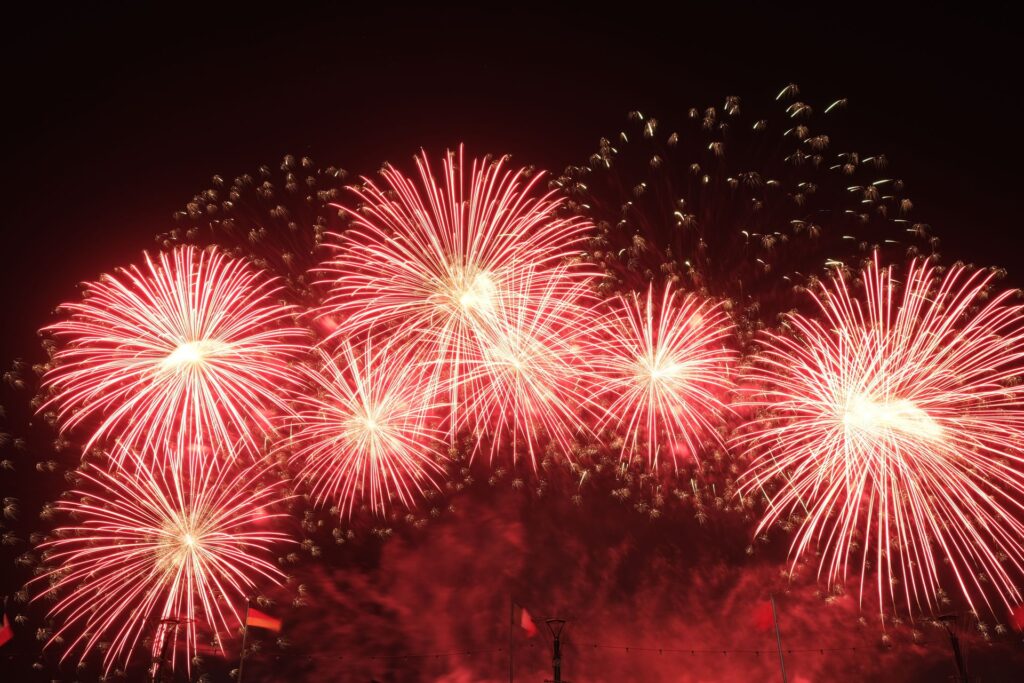 Multiple bright red fireworks fill the night sky with dazzling bursts of light during a large fireworks display.