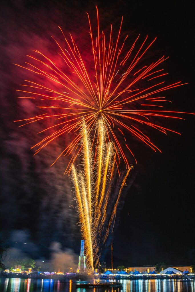 A large red firework explodes above golden sparkling trails, lighting up the night sky over a waterfront festival.