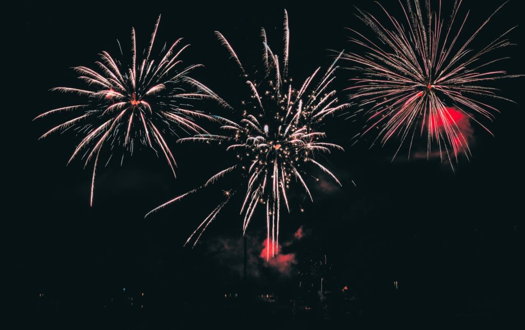 Three white and pink fireworks burst across a dark night sky, creating delicate trails of sparkling light.