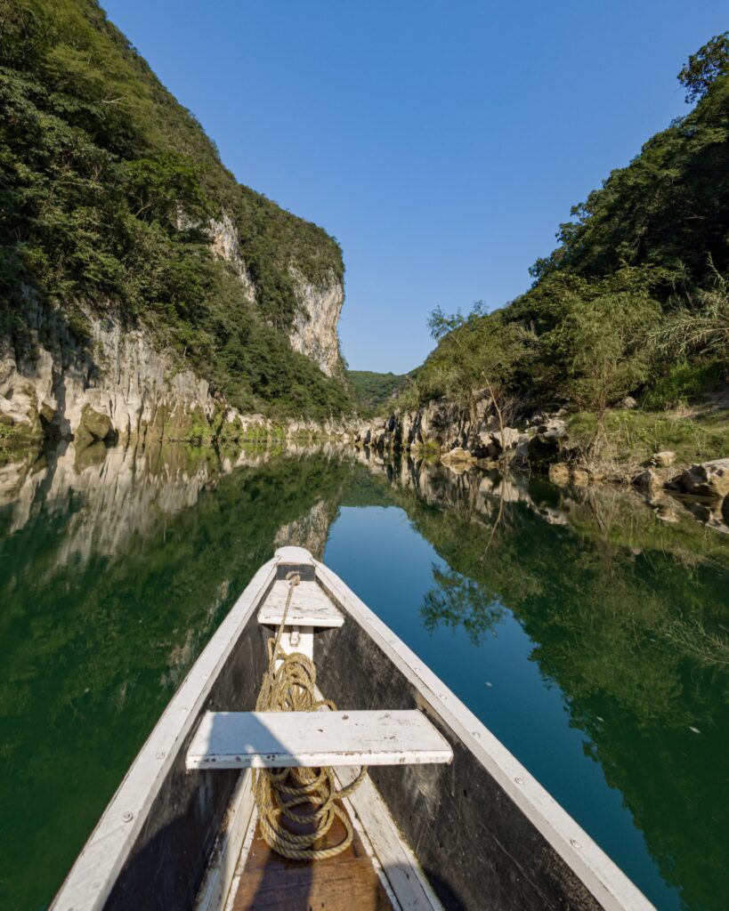 Boat traveling through a peaceful river gorge surrounded by towering rocky cliffs and lush green forests, with reflections on the calm water.