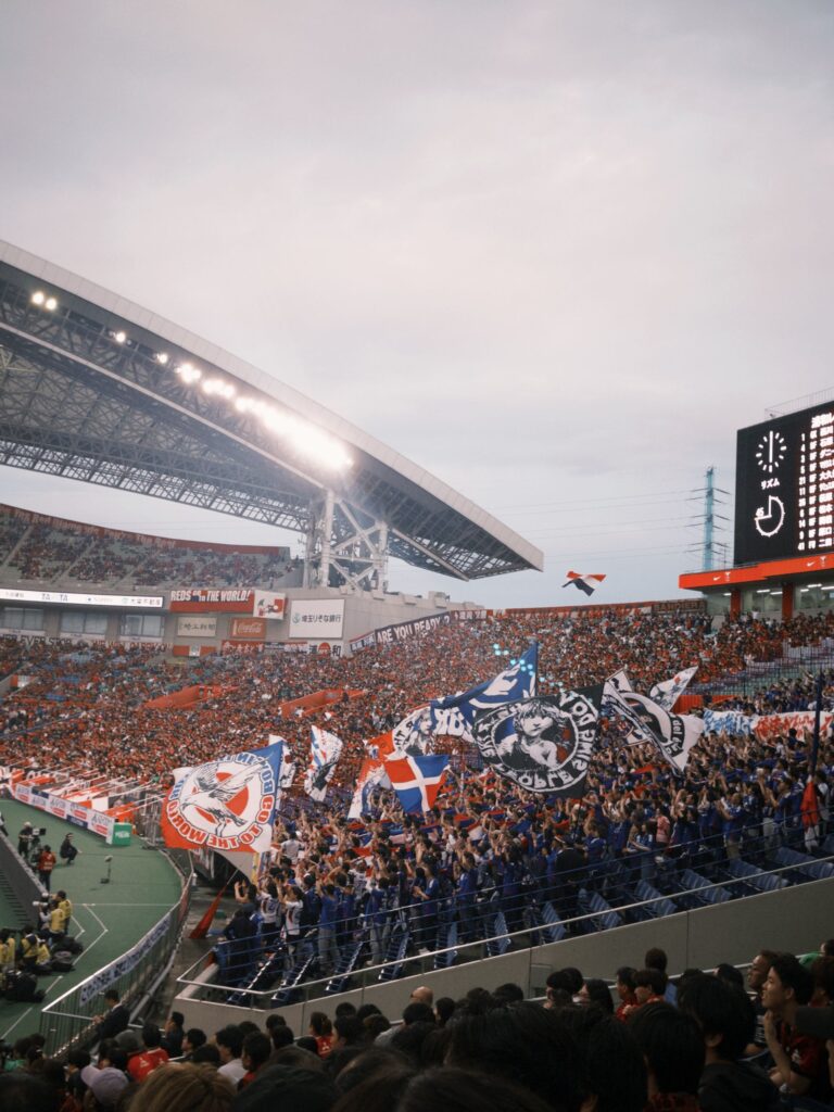 Football supporters waving colorful flags and cheering inside a packed stadium during a match involving the Urawa Reds in Saitama.