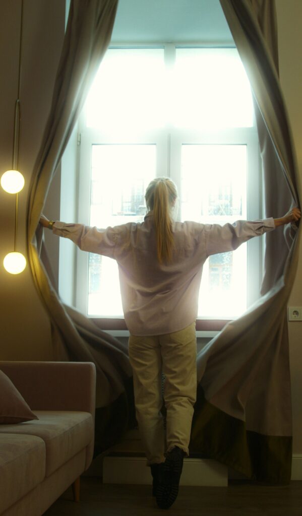 A woman opens thick curtains to let natural daylight into a room, showing how curtains can help regulate warmth in a home during winter.